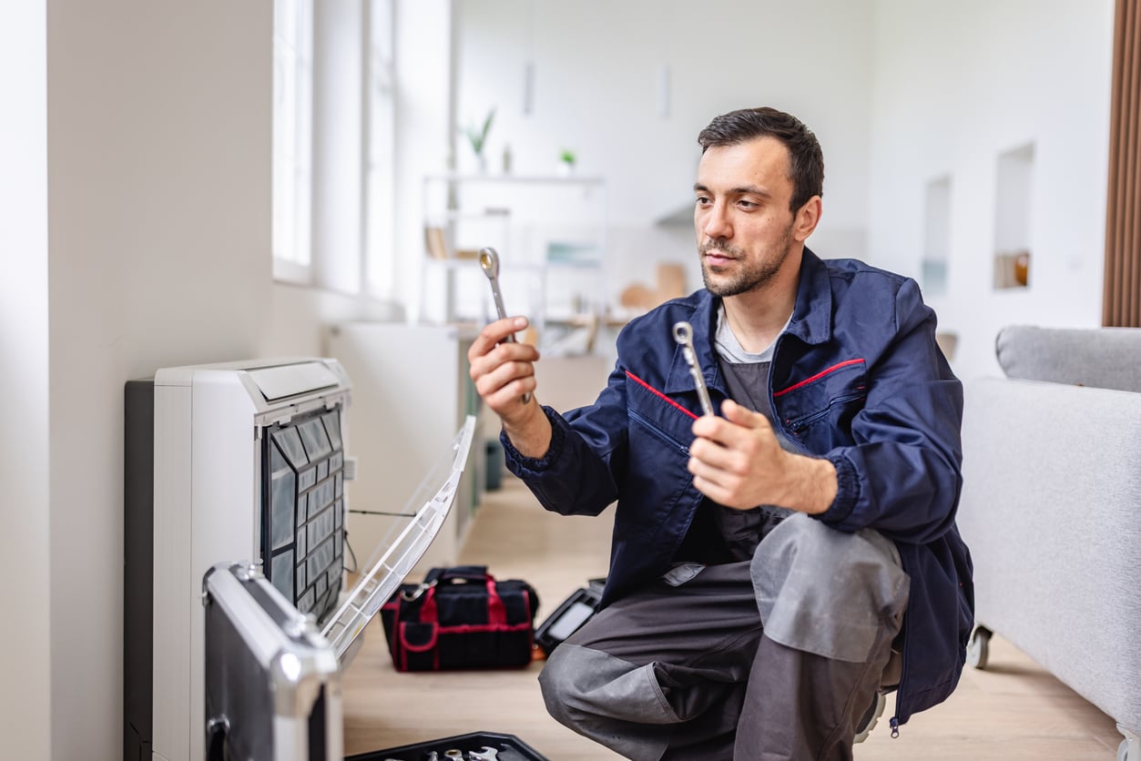 A repairman in work clothes kneels by an open heating unit, showcasing his DIY skills as he examines two wrenches. A toolbox and tools are scattered on the floor in a modern, bright living room.