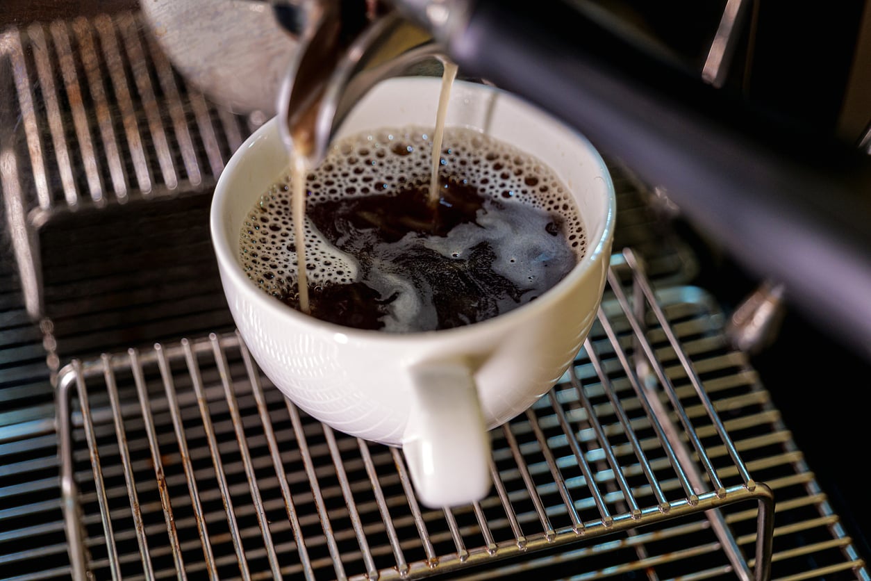 A close-up of a white cup being filled with fresh coffee from an espresso machine, sitting on a metal rack.