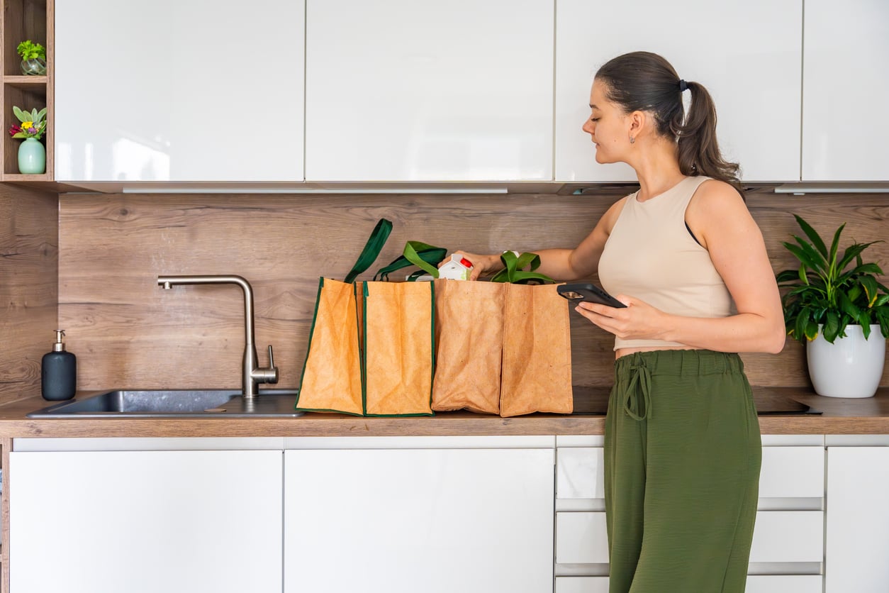 A woman in a beige tank top and green pants unpacks groceries from reusable bags on a kitchen counter while holding her phone, perhaps comparing 1991 prices to today’s. The modern kitchen features white cabinets, a plant, and a wooden backsplash.