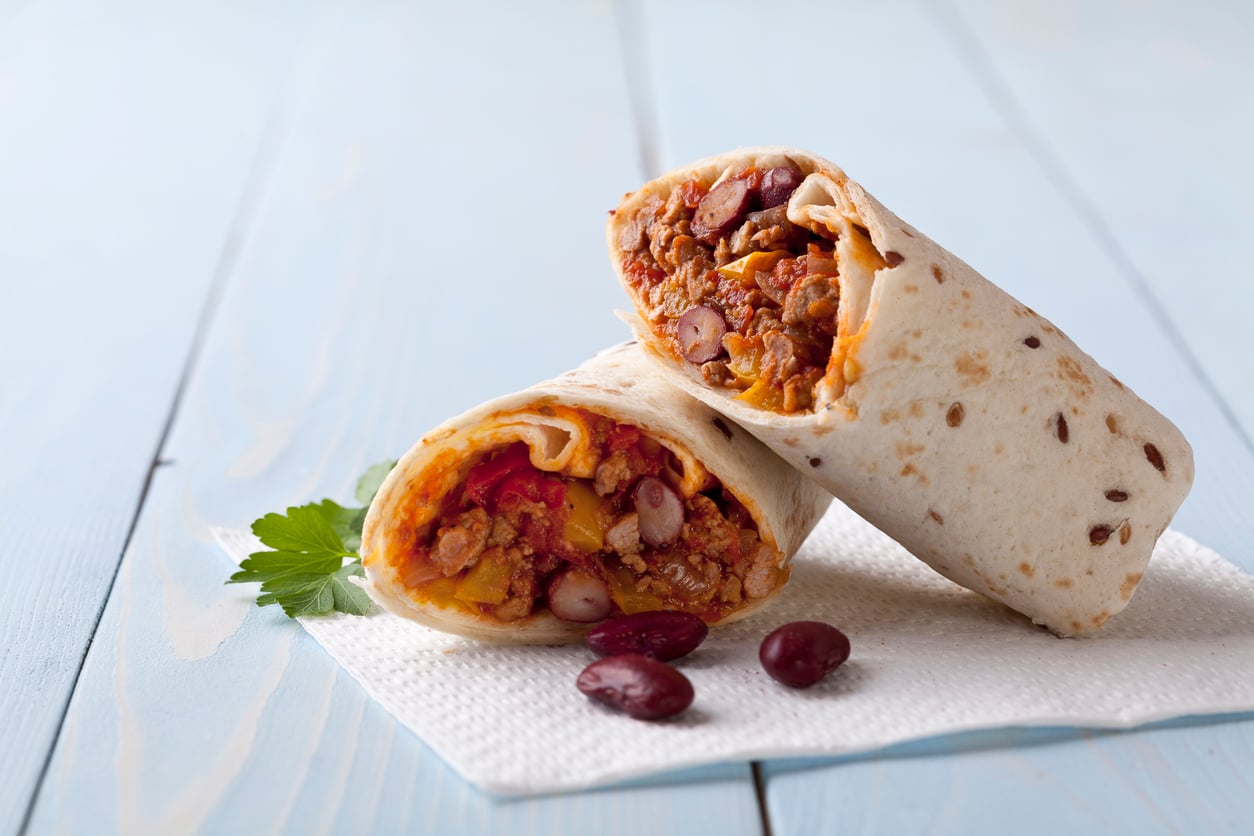 Two burritos filled with ground meat, beans, vegetables, and egg substitutes are stacked on a white napkin on a light wooden surface, with parsley and a few red kidney beans beside them.