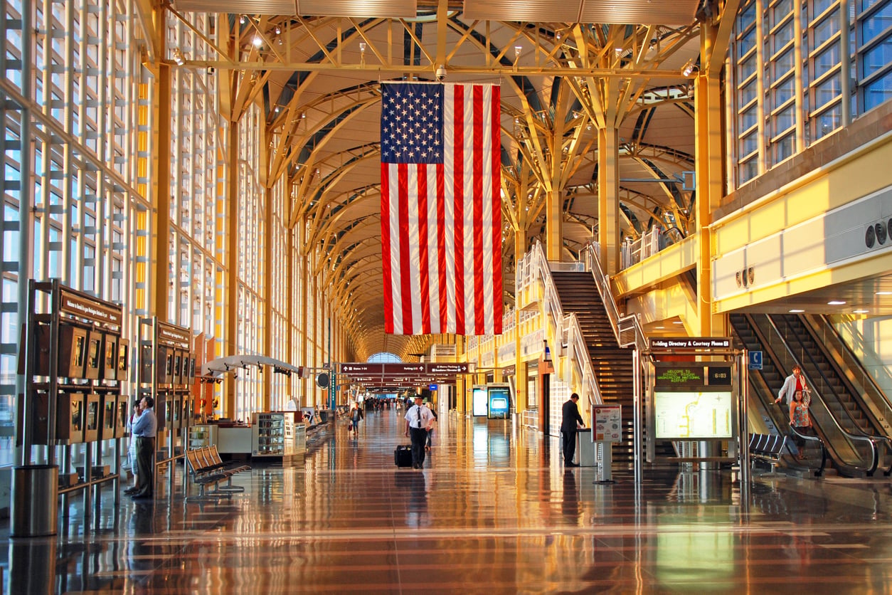 A spacious, convenient airport terminal with high arched ceilings, large windows, and an American flag hanging in the center. Travelers walk along the polished floor, with clear signage and escalators enhancing the convenience for all.