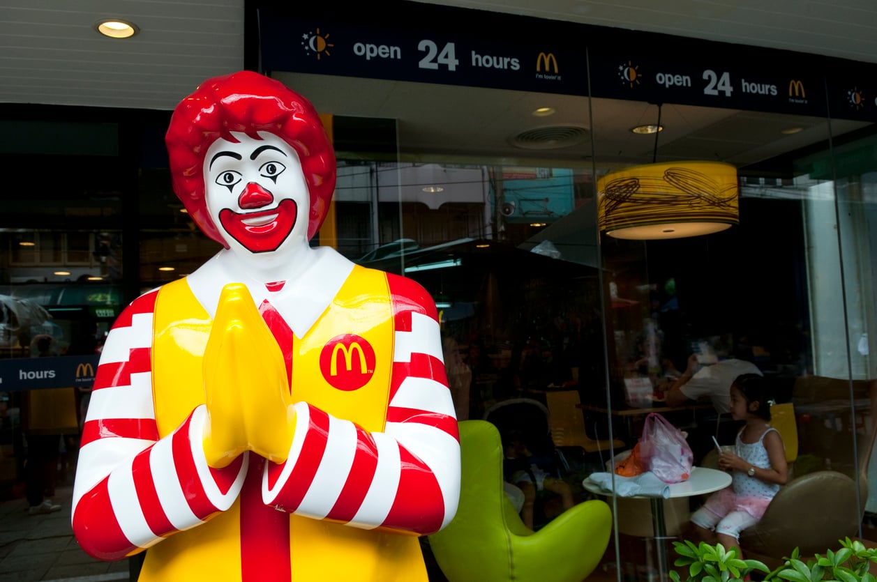 A statue of Ronald McDonald with hands pressed together in a greeting stands outside one of the Coolest McDonald's Locations, marked by a sign that says "open 24 hours." People are seated inside the restaurant.