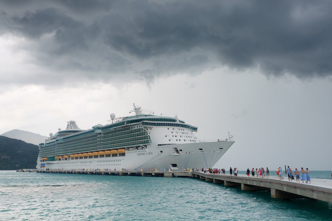 A large white cruise ship is docked at a pier with people walking along it. The sky is dark and cloudy, suggesting an approaching storm, and mountains are visible in the background—perfect for testing your favorite travel hacks.