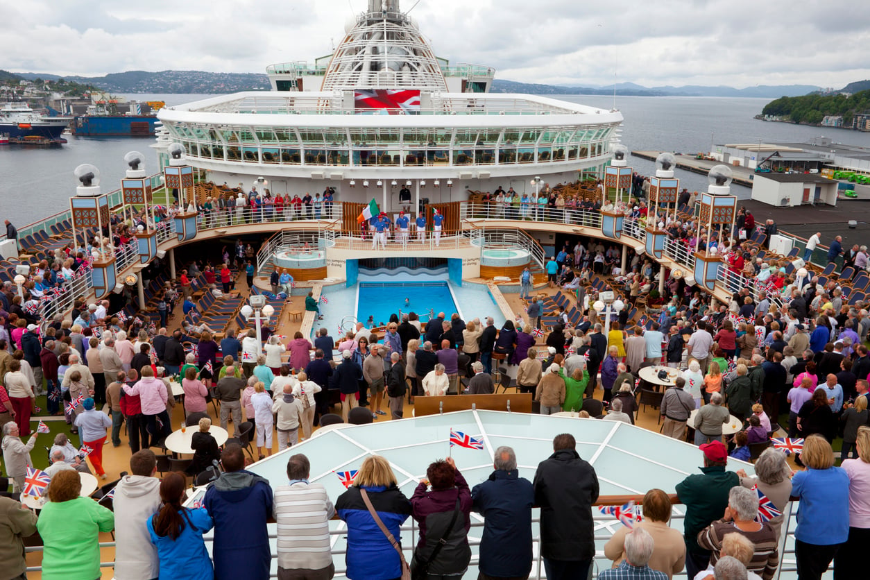 A large crowd gathers around a swimming pool on the Best Cruise Line 2026 cruise ship, watching an event. British flags wave as the ship is docked near a scenic shoreline with hills and water beneath a cloudy sky.