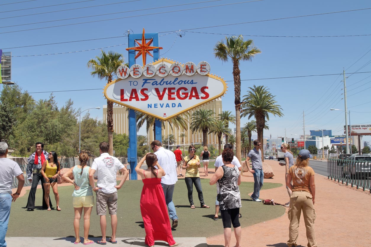A group of people stands in front of the famous "Welcome to Fabulous Las Vegas, Nevada" sign on a sunny day, ready to explore the city and share their favorite Vegas money-saving tips amid palm trees and city buildings.