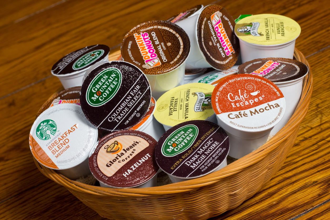 A wicker basket filled with assorted single-serve coffee pods from various brands, including a selection of sometimes overpriced products like Starbucks, Green Mountain, Gloria Jean’s, Dunkin’, and Café Escapes, sits on a wooden surface.