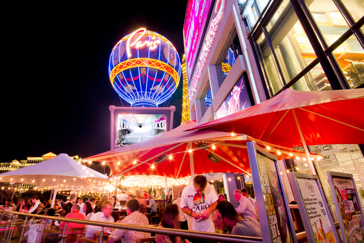 Outdoor dining area at night with people seated under red umbrellas, string lights above, and the illuminated Paris Las Vegas balloon sign glowing brightly in the background—a perfect spot to enjoy the scene while discovering Vegas money-saving tips.