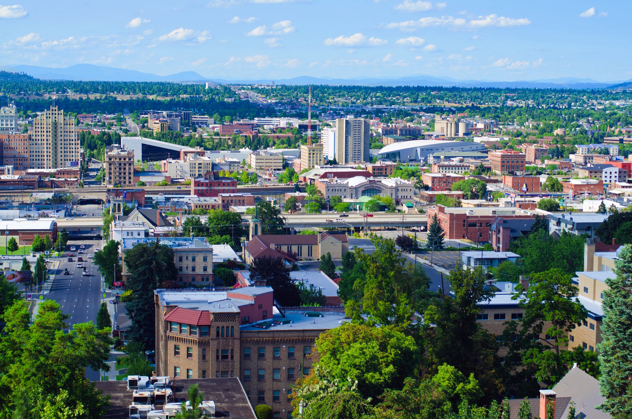 A cityscape featuring a mix of modern and older buildings surrounded by trees, with mountains visible in the background under a bright, partly cloudy sky.