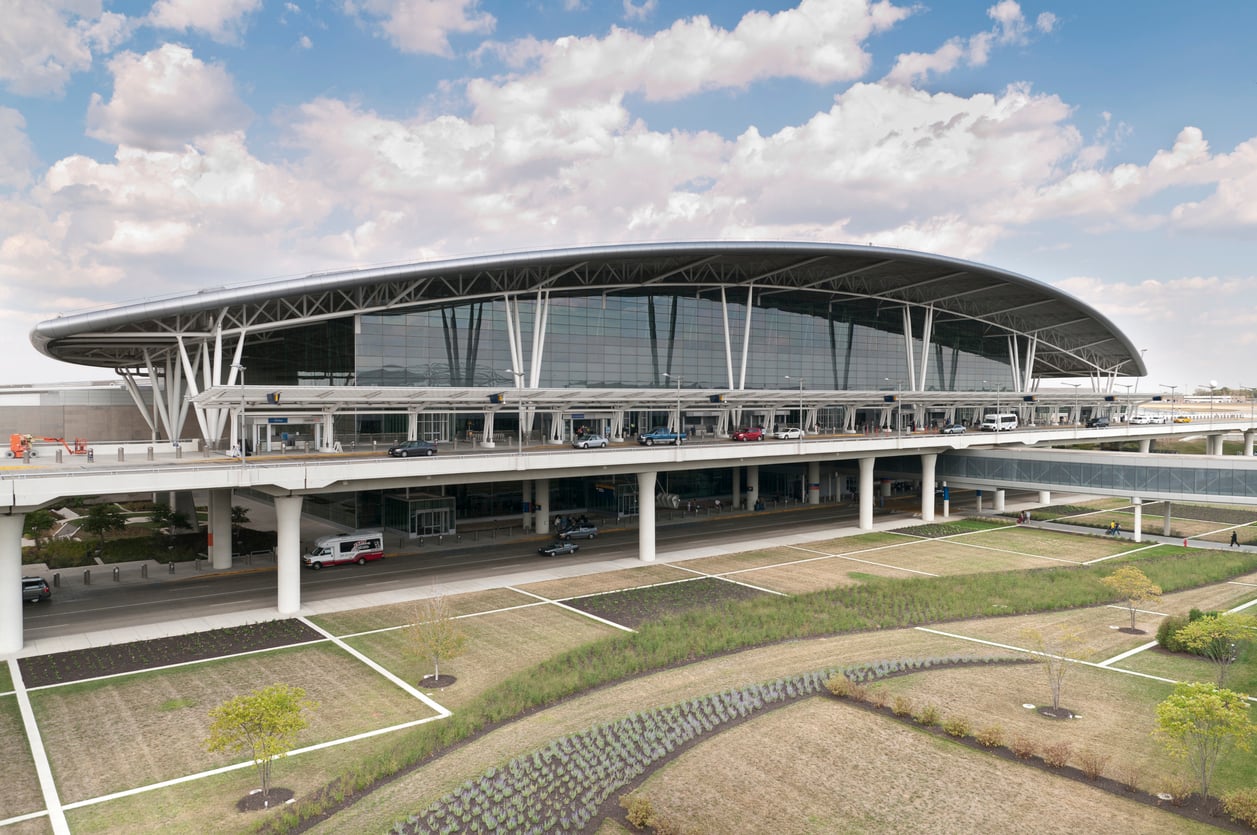 A modern airport terminal with a curved roof, large glass windows, and elevated roadways offers convenient airports access, surrounded by landscaped grounds and cloudy blue skies.