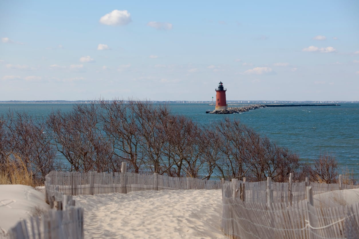 A sandy path lined with a wooden fence leads to the beach, with leafless trees on either side and a red lighthouse standing on a rocky jetty in the distance against the blue sea and sky.