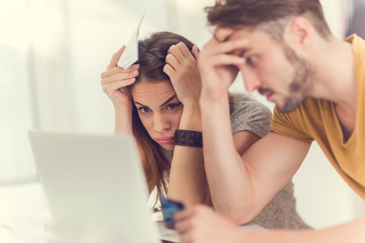 A woman and man sit in front of a laptop looking stressed. The woman holds a piece of paper to her forehead while the man touches his head, both appearing worried or frustrated.