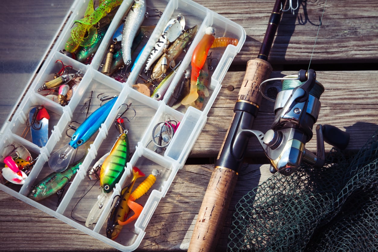 A tackle box with assorted fishing lures and hooks sits on a wooden surface next to a fishing rod, reel, and green fishing net—timeless products men still buy for their weekend angling adventures.