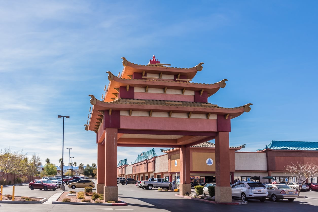 A large decorative gateway with traditional East Asian architectural style stands in front of a shopping plaza, a popular spot for those seeking Las Vegas budget tips, surrounded by parked cars under a clear blue sky.