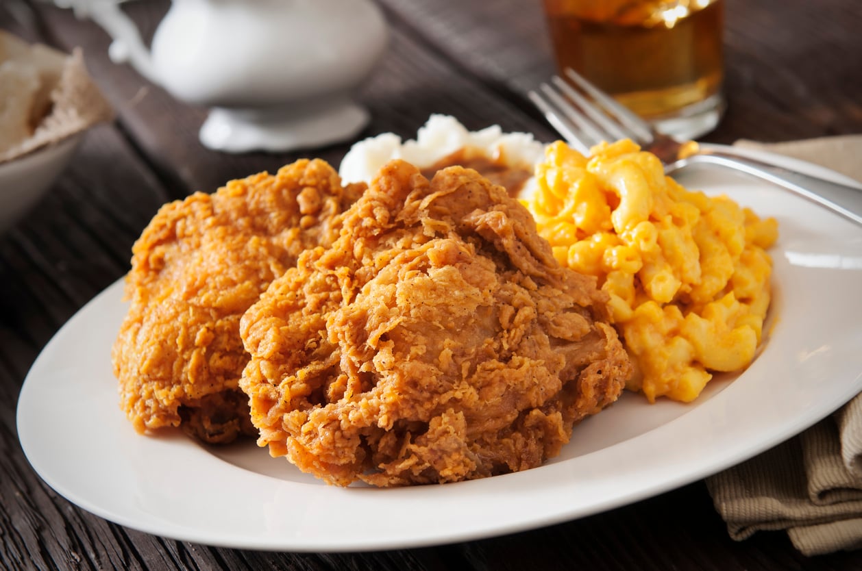 A plate of crispy fried chicken with a serving of creamy macaroni and cheese and mashed potatoes, set on a rustic wooden table with a fork and knife beside the plate.