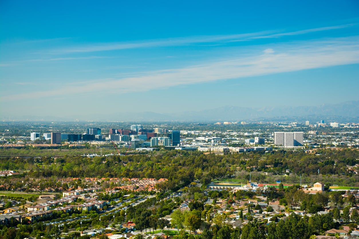 Aerial view of a cityscape with a mix of low-rise houses, green trees, and tall modern buildings under a clear blue sky with distant mountains in the background.