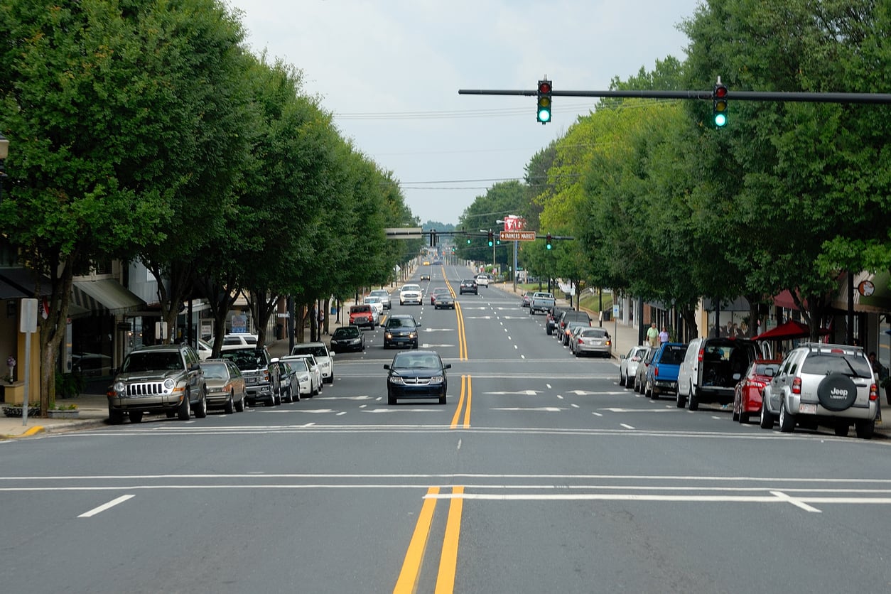 A straight, tree-lined city street with cars parked on both sides and vehicles driving in both directions under green traffic lights on a cloudy day. Buildings and shops are visible along the sidewalks.
