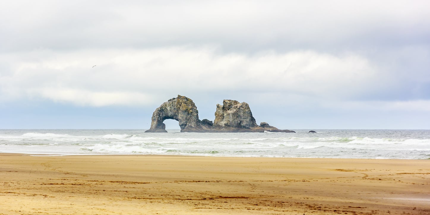 A sandy beach stretches toward the ocean, where a large rock formation with a natural arch stands amid the waves under a cloudy sky—one of many scenic views found near charming beach towns.