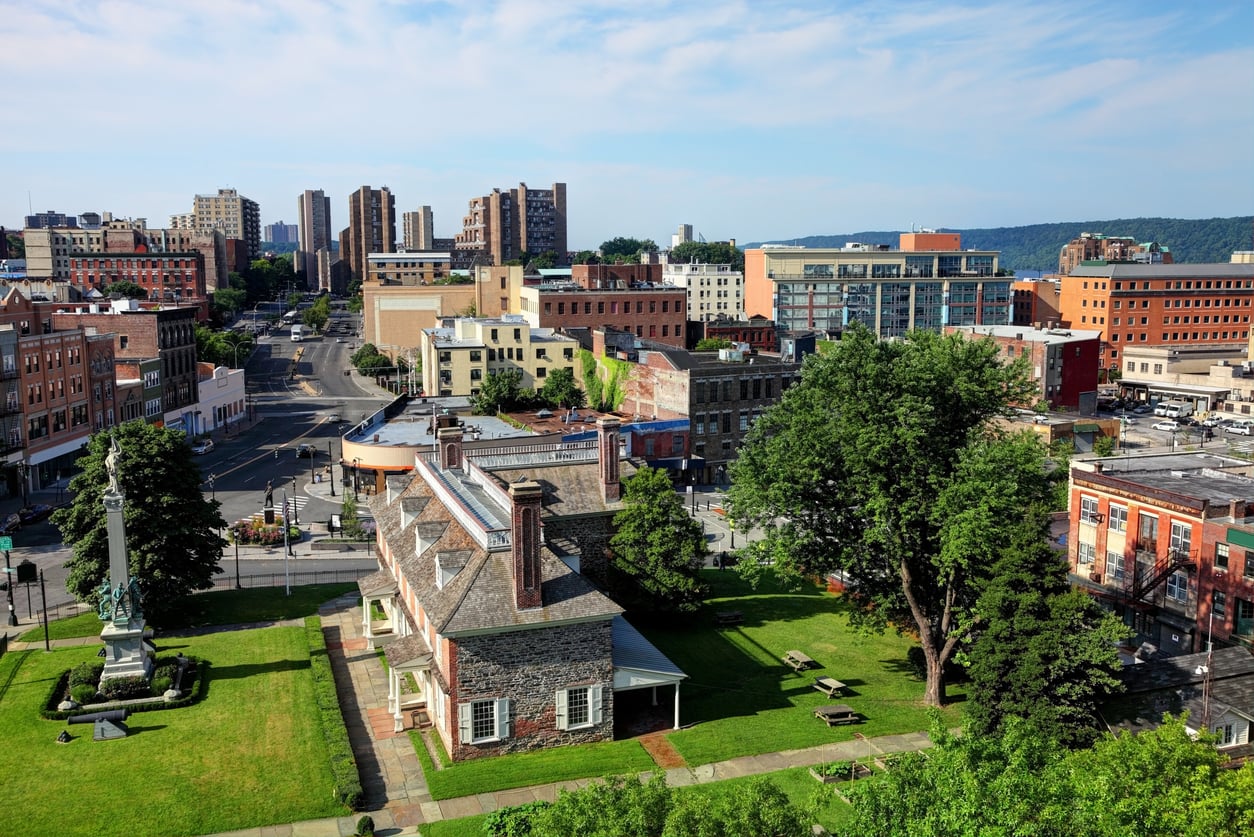 A historic house with a large green lawn sits in the foreground, surrounded by trees, with mid-rise buildings, busy streets, and a city skyline in the background under a blue sky.