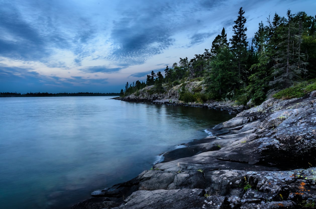 Rocky shoreline with evergreen trees beside a calm lake under a cloudy blue sky at dusk. The water is still, and the landscape appears serene and untouched by humans.
