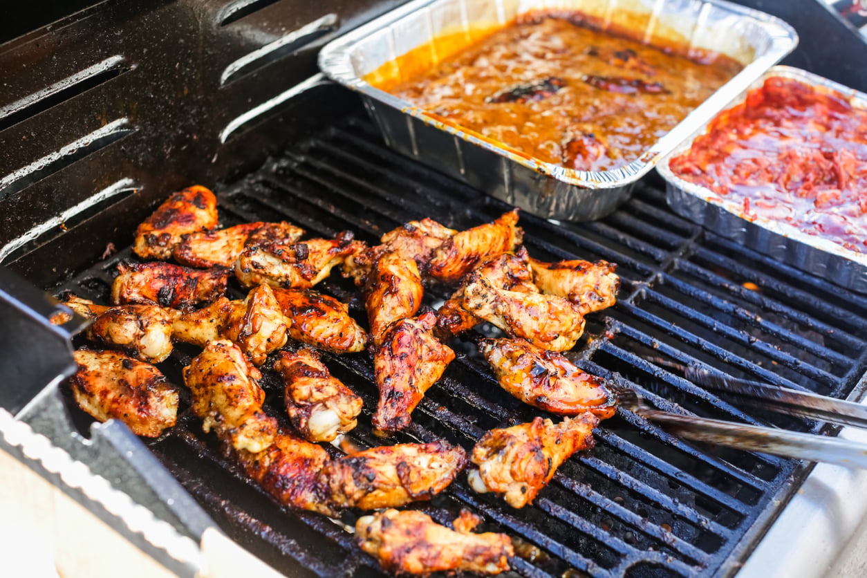 Grilled chicken wings cooking on a barbecue grill, with metal tongs and two foil trays containing sauces or side dishes placed on the grill beside the chicken.