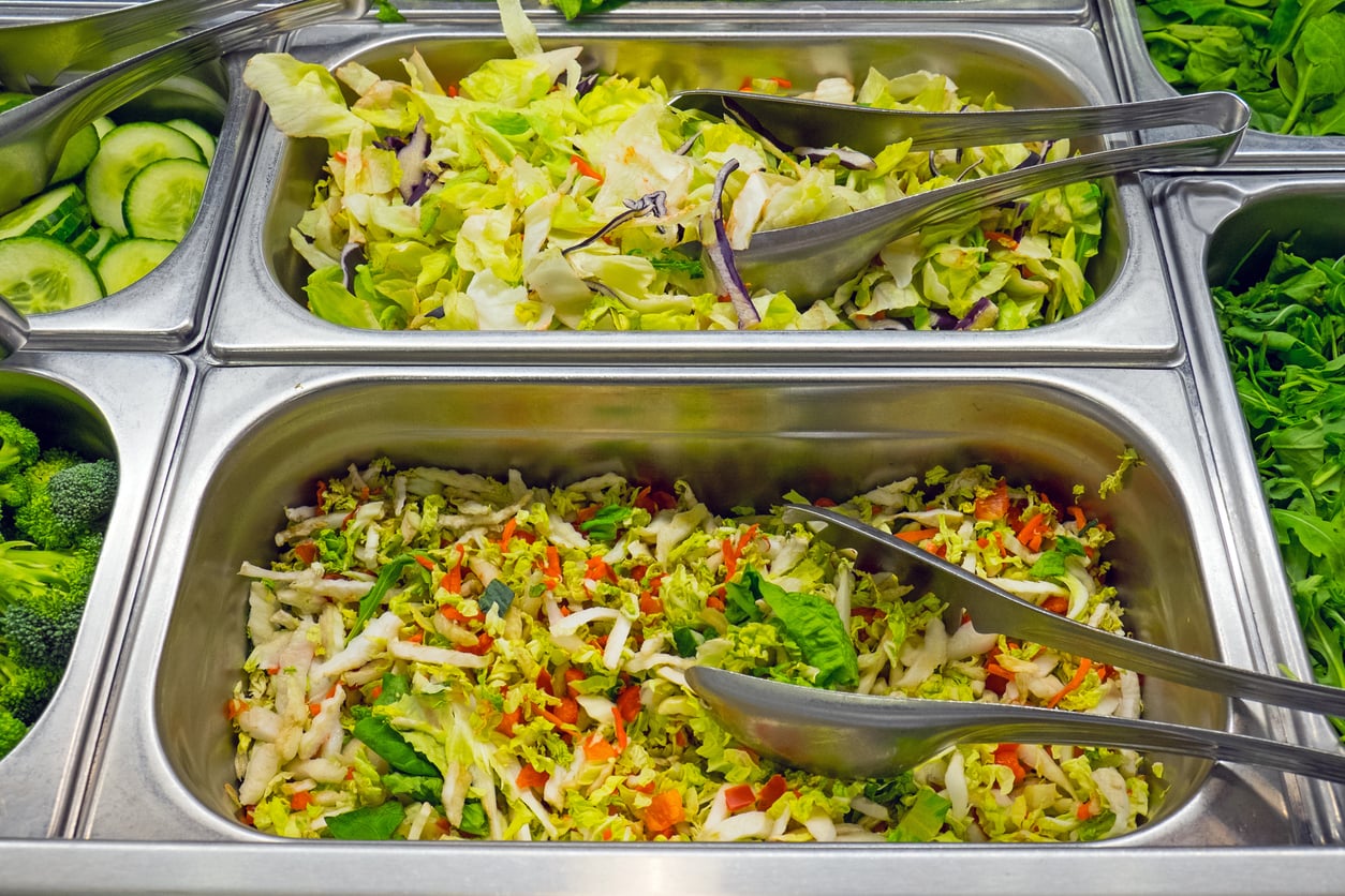 Two metal trays in a salad bar, one filled with shredded lettuce and cabbage mix, the other with chopped mixed vegetables, both with metal tongs. Surrounding trays hold cucumbers, broccoli, and leafy greens.