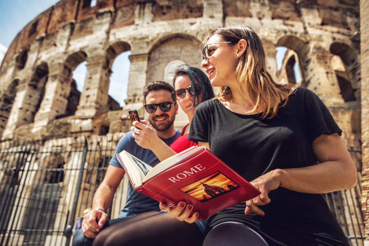 Three people sit in front of the Colosseum in Rome. One woman holds a guidebook titled "ROME," while the others look at a mobile phone and smile, enjoying their sightseeing experience.