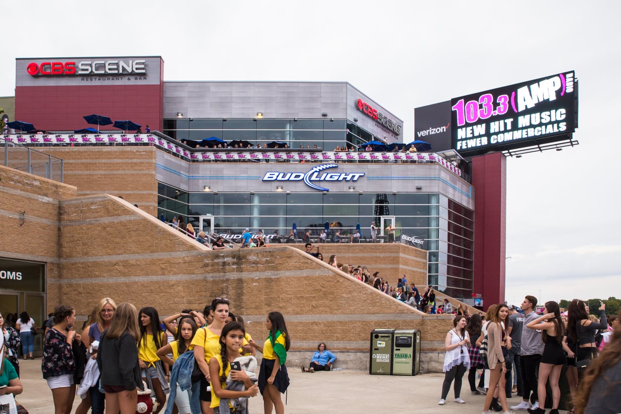 A crowd of people stands and walks near a large brick building with “CBS Scene” and “Bud Light” signs. Above the venue, a digital billboard advertises 103.3 AMP radio and Verizon—perhaps the perfect spot to share concert ticket hacks.