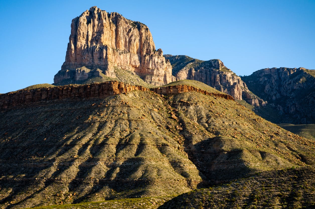 A dramatic, sunlit mesa with steep, rugged slopes rises prominently against a clear blue sky, casting long shadows across the rocky landscape. Sparse vegetation covers the arid ground.