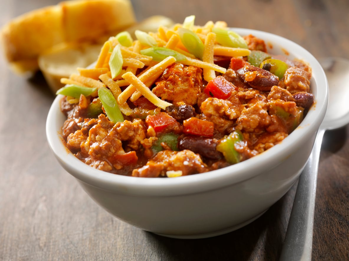 A bowl of chili topped with shredded cheddar cheese and sliced green onions, featuring ground beef alternatives, beans, and diced vegetables, with a piece of bread in the background.