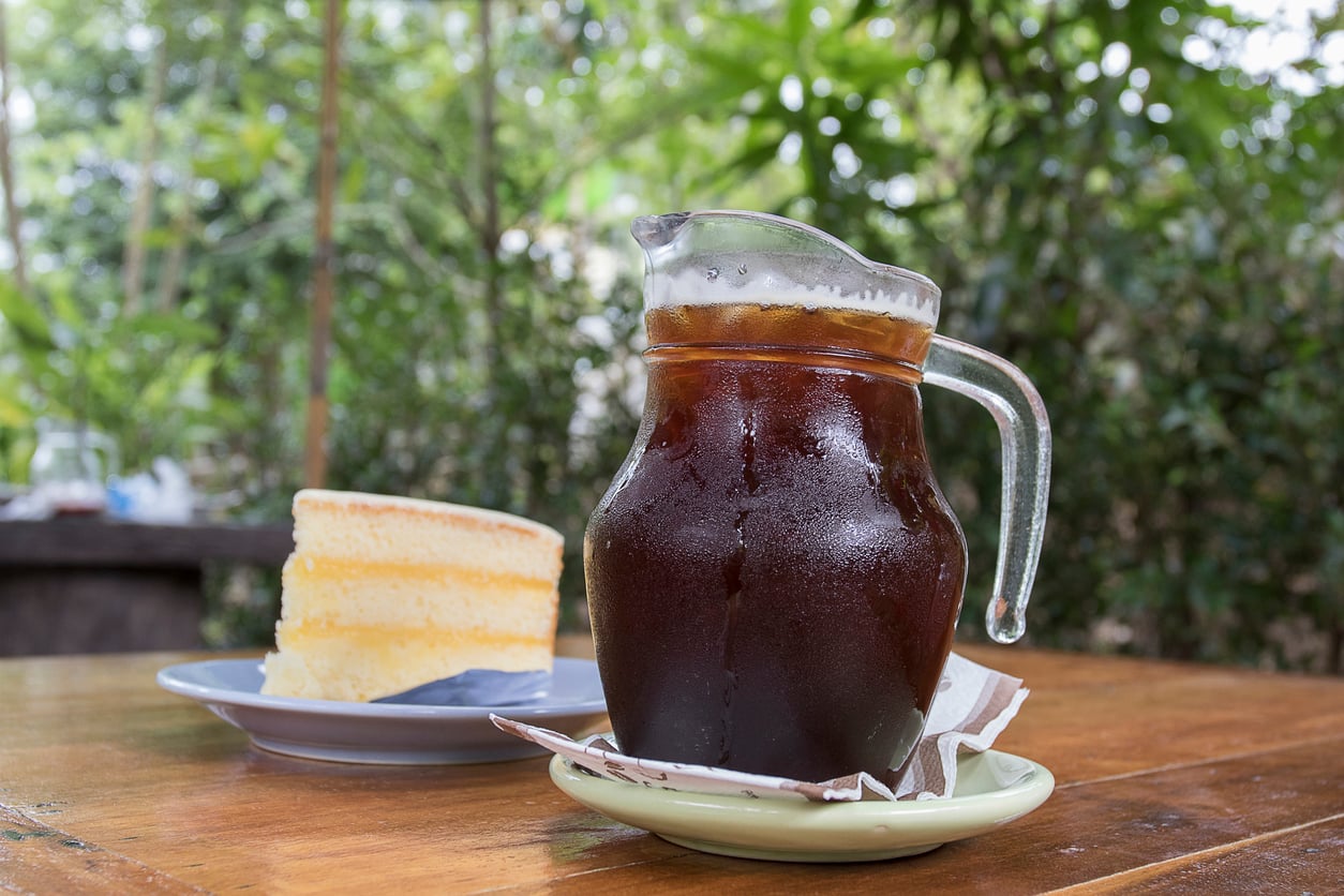 A glass pitcher of iced coffee sits on a wooden table next to a plate with a slice of layered yellow cake, with a green, leafy outdoor background.
