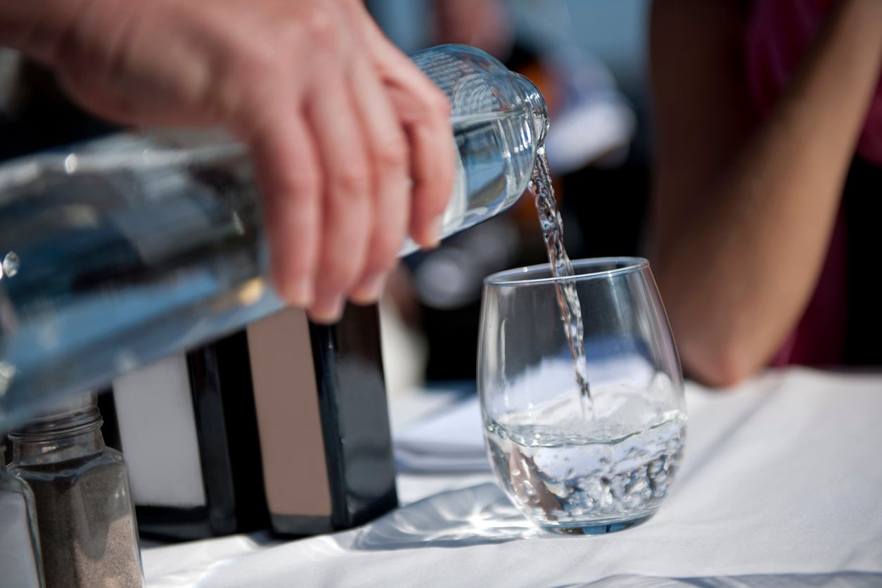 A person pours water from a glass bottle into a glass on a white tablecloth, with salt and pepper shakers and another person sitting nearby in the background.