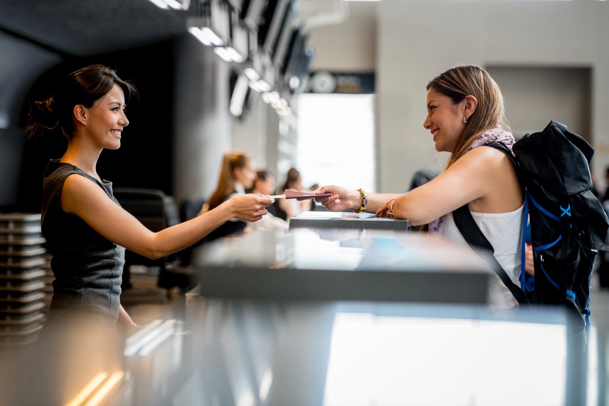 A smiling airline agent hands a boarding pass to a traveler with a backpack at an airport check-in counter. Both women are facing each other and appear friendly and engaged.