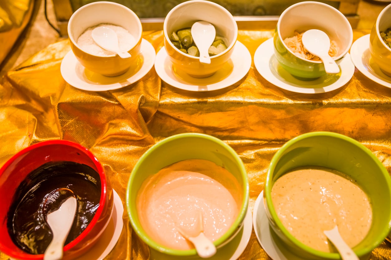 Six bowls of various sauces or condiments are arranged on a golden cloth, each with a white spoon. The bowls contain different colored spreads, possibly for a buffet or self-serve station.