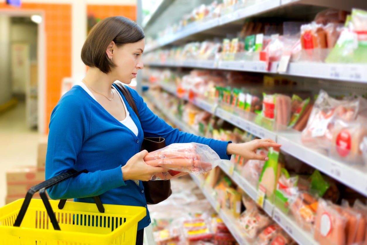 A woman in a blue sweater holds a shopping basket and packaged sausages, examining ground beef alternatives on a refrigerated grocery store shelf.