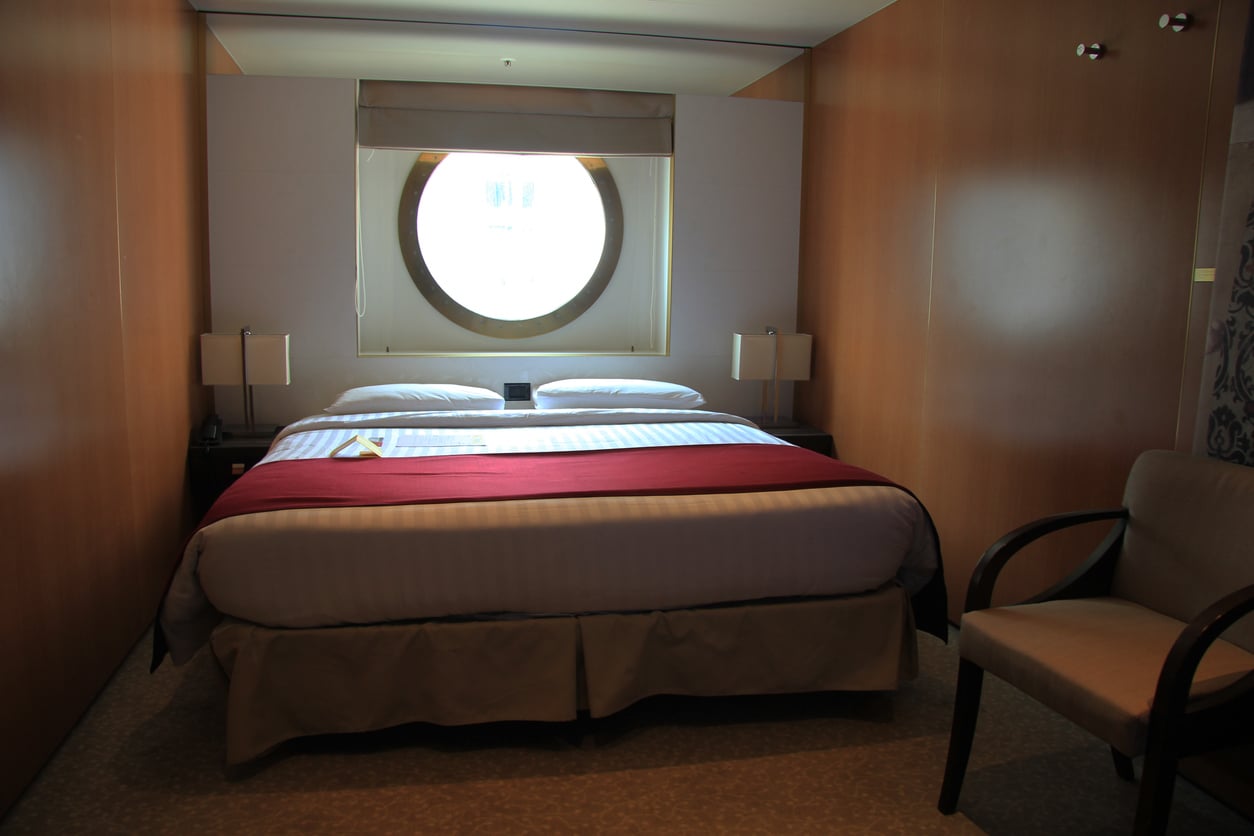 A neatly made bed with a red runner is centered in a cozy room with wood-paneled walls and a circular porthole window above the headboard—a perfect setting to relax and unwind after trying out your favorite travel hacks. Two lamps and a chair are nearby.