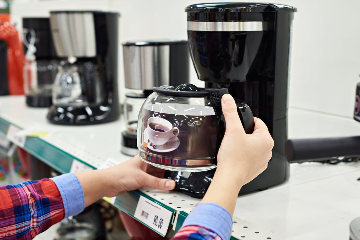A person in a plaid shirt holds a glass coffee pot in front of a black coffee maker on a store shelf. The pot has an image of a cup of coffee printed on it, and price tags are visible nearby.