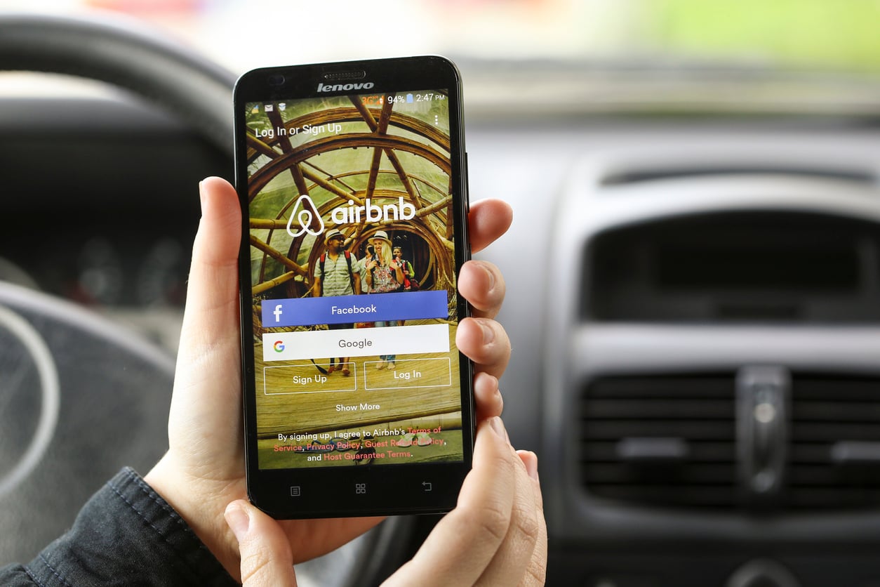 A person holds a Lenovo smartphone displaying the Airbnb login screen with options to log in via Facebook, Google, or email. The phone is held inside a car, with the dashboard visible in the background.