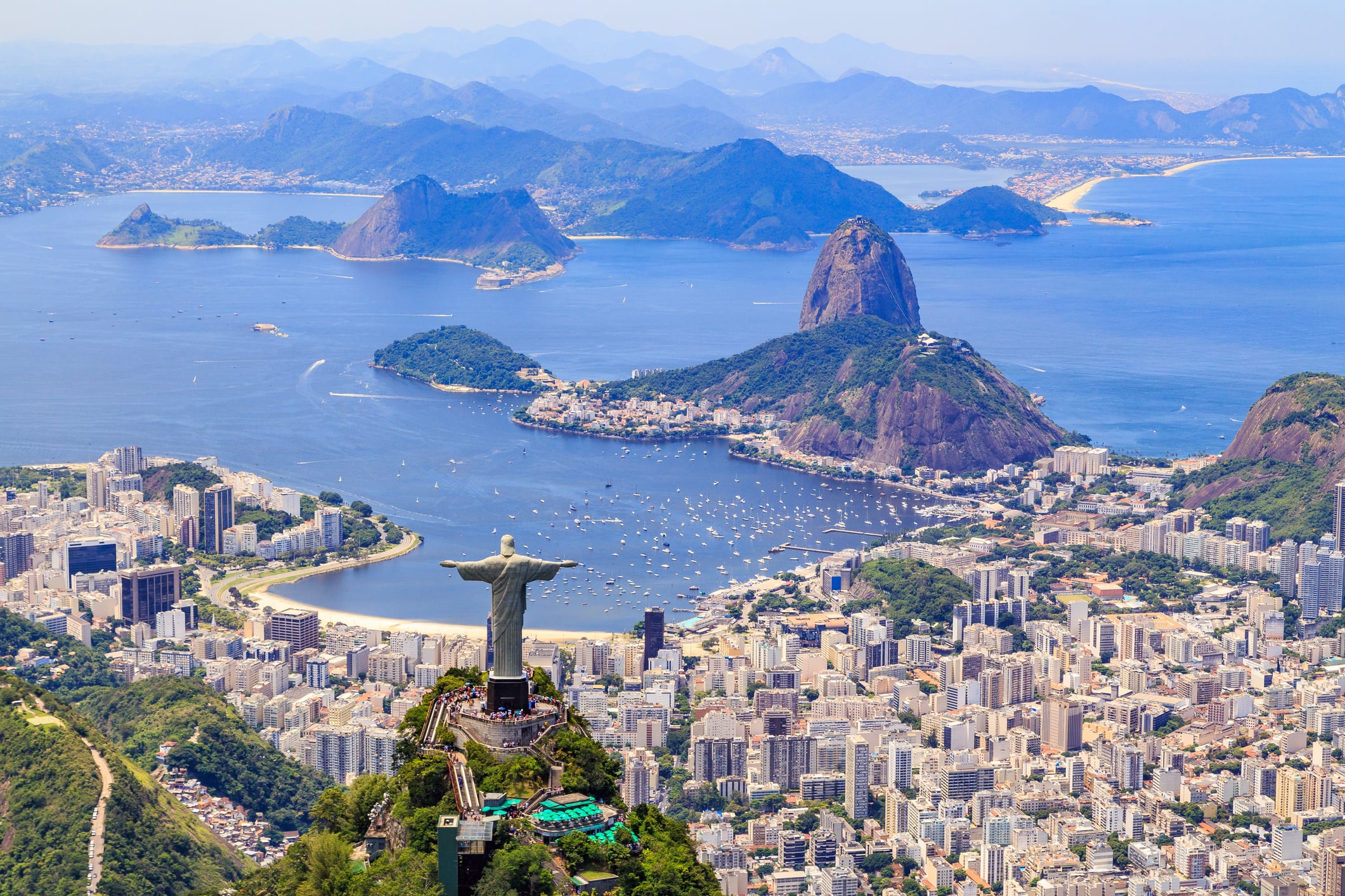 Aerial view of Rio de Janeiro featuring the Christ the Redeemer statue, Sugarloaf Mountain, city buildings, and Guanabara Bay with boats, set against distant mountains under a clear blue sky.