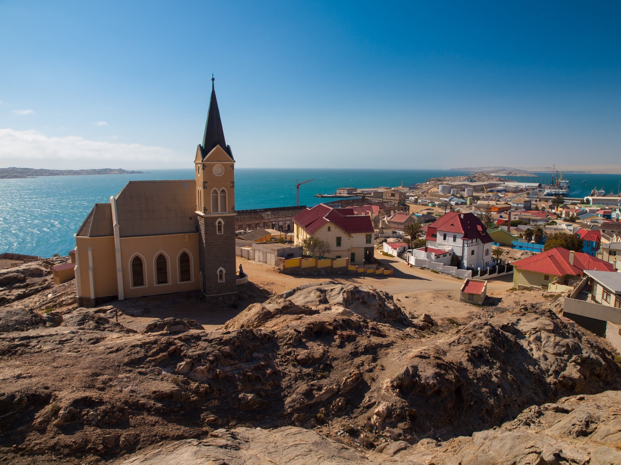 A church with a tall steeple stands on rocky terrain overlooking a coastal town with colorful rooftops, near a bright blue sea under a clear sky.