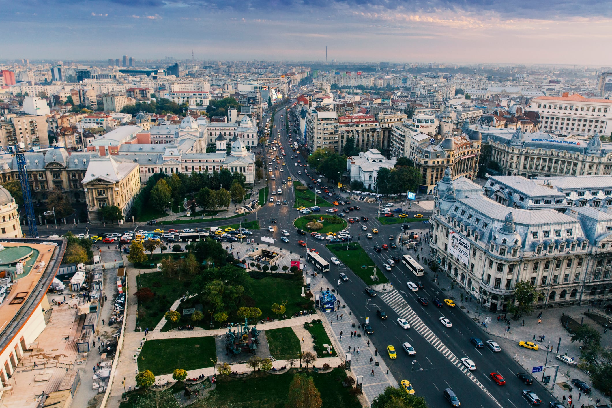 Aerial view of a busy city intersection with historic buildings, green parks, and multiple streets filled with cars and people under a partly cloudy sky.