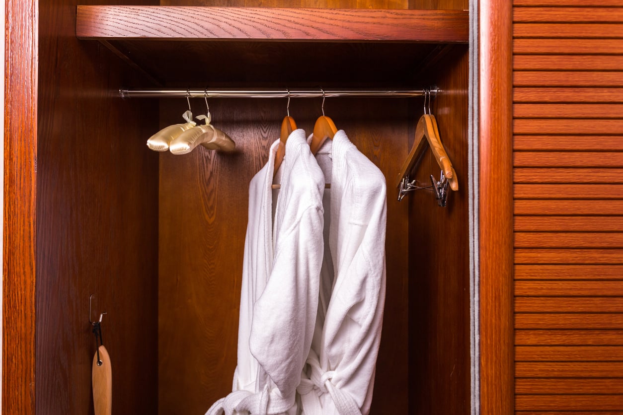 Two white bathrobes hang on wooden hangers inside a wooden wardrobe. There are additional hangers and a shoehorn also visible inside the wardrobe.