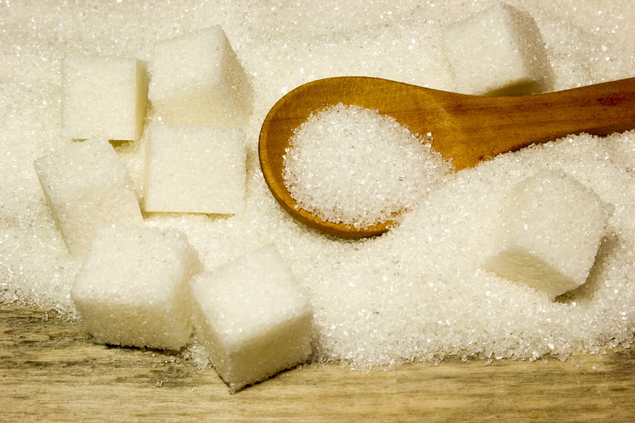 A wooden spoon with granulated sugar rests on a pile of sugar, next to several white sugar cubes on a wooden surface.