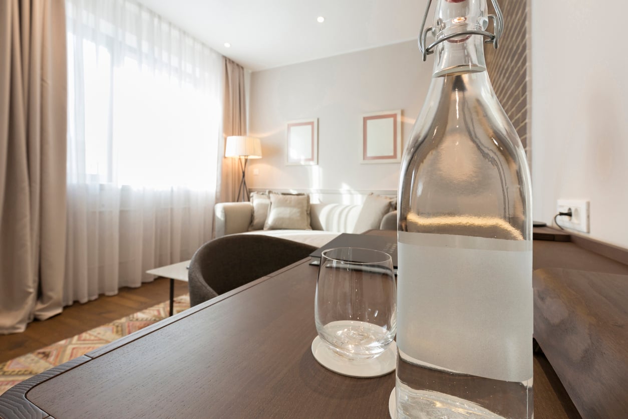 A clear glass bottle of water and an empty glass sit on a wooden table in a bright, modern hotel room with a beige sofa, framed art, and a large window covered by sheer curtains—emphasizing both comfort and hotel room safety.