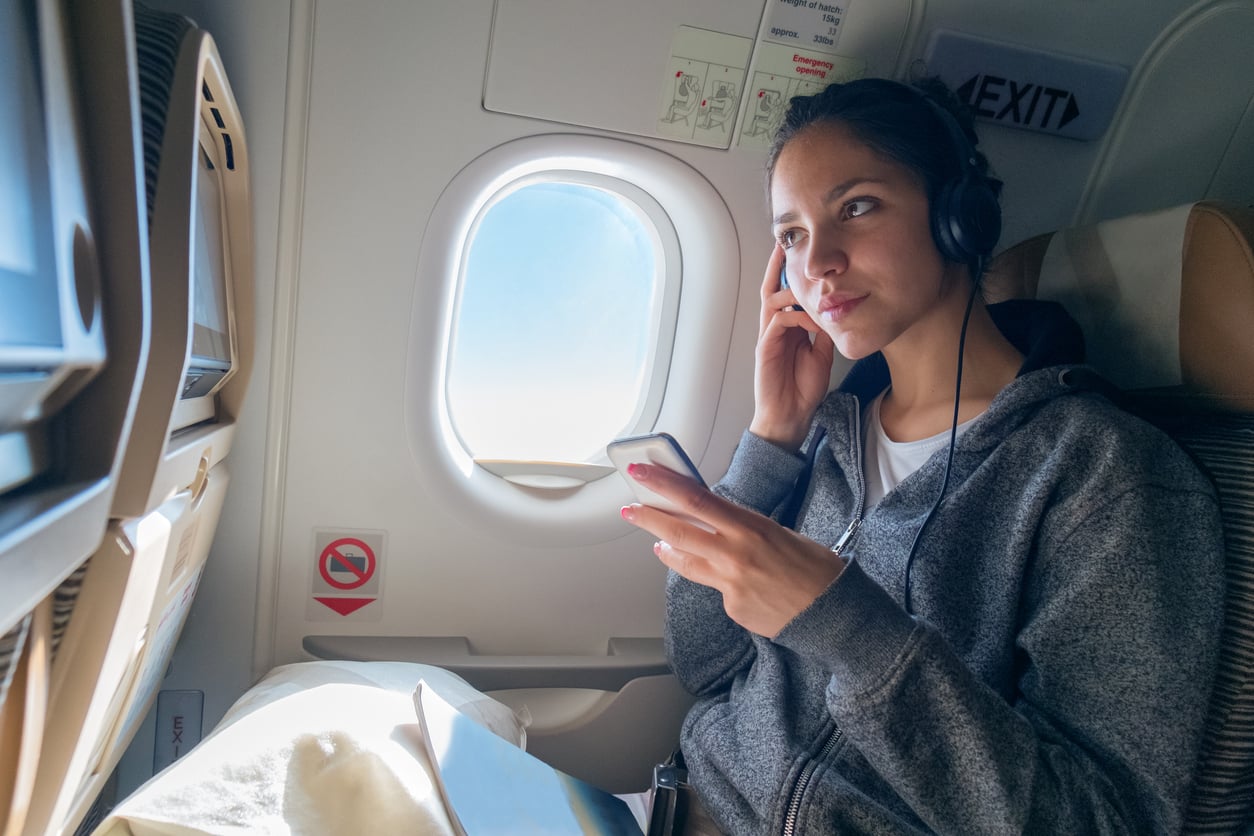 A woman sitting by an airplane window wears headphones and holds a smartphone, looking thoughtful. Sunlight streams through the window, illuminating her face. She has a laptop and a blanket on her lap.