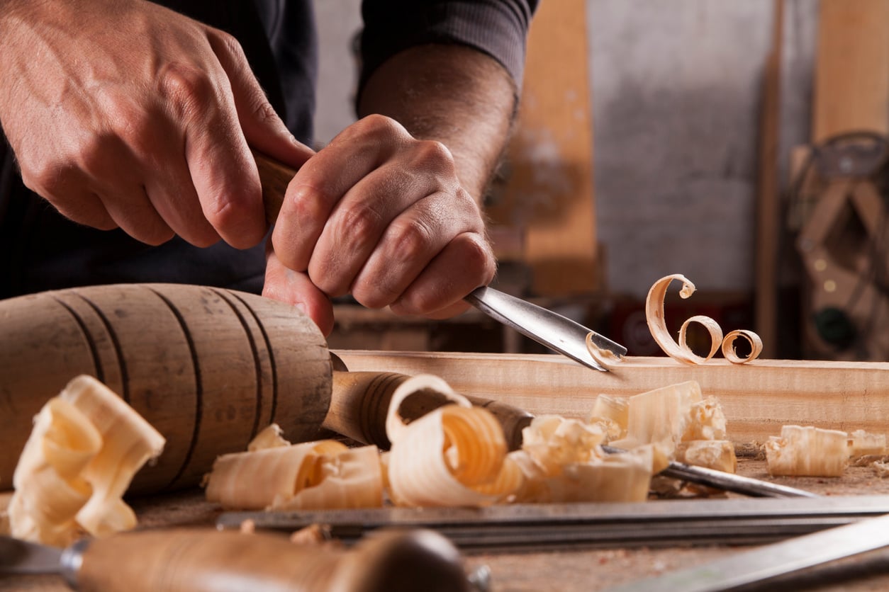 A person uses a chisel and mallet to carve wood, creating curly wood shavings. Scattered tools hint at detailed woodworking in progress—a great example of budget-friendly hobbies for creative hands.