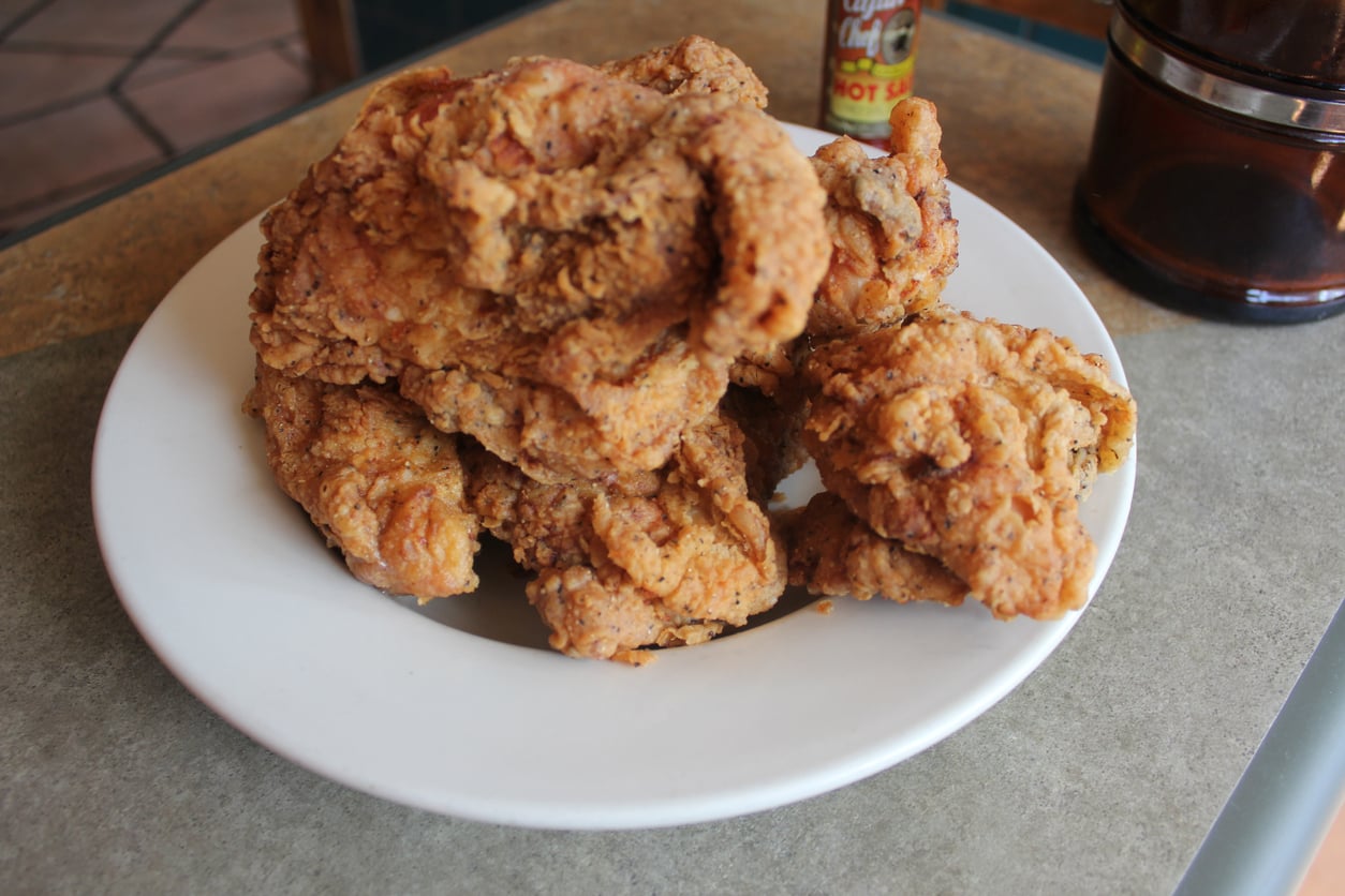 A plate with several pieces of crispy fried chicken sits on a table. A bottle of hot sauce and part of a brown container are visible in the background.