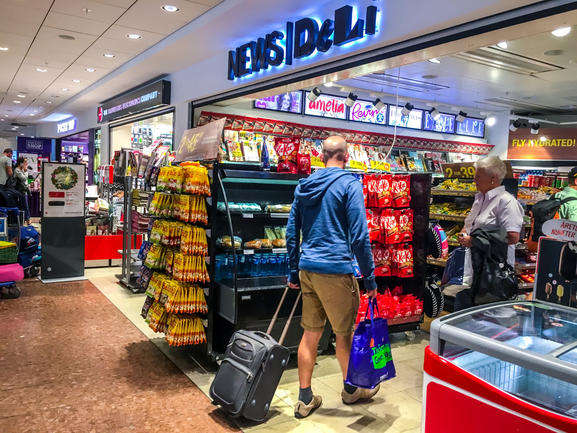 A man pulls a suitcase past a "News Deli" shop filled with snacks. He holds a blue shopping bag and wears a blue hoodie and khaki shorts.