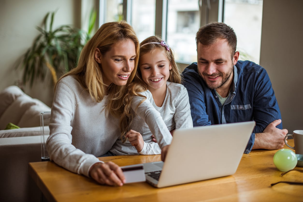 A smiling woman, man, and young girl sit together at a table, looking at a laptop screen. The woman holds a credit card while the girl points excitedly—perhaps they're searching for theme park hacks to plan their next adventure online at home.