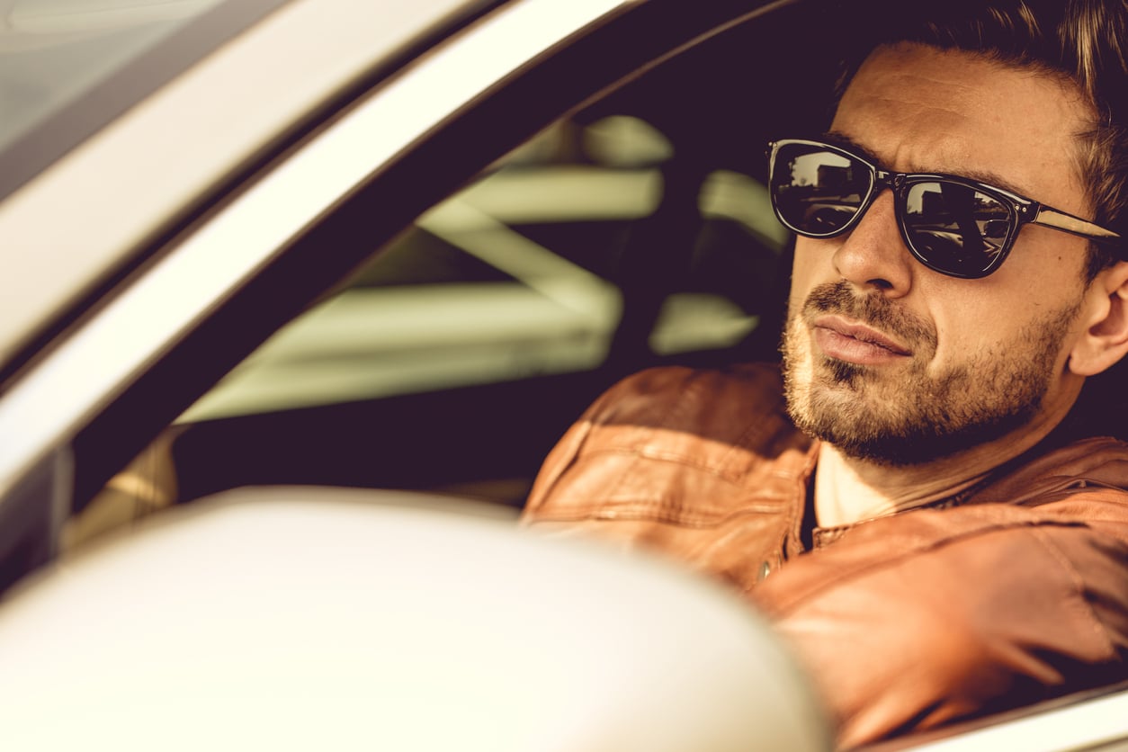 A man with a beard wearing sunglasses and a brown leather jacket sits in the driver's seat of a car, embodying the confidence often seen in ads for products men skip buying, as he looks out the window with an assured expression.