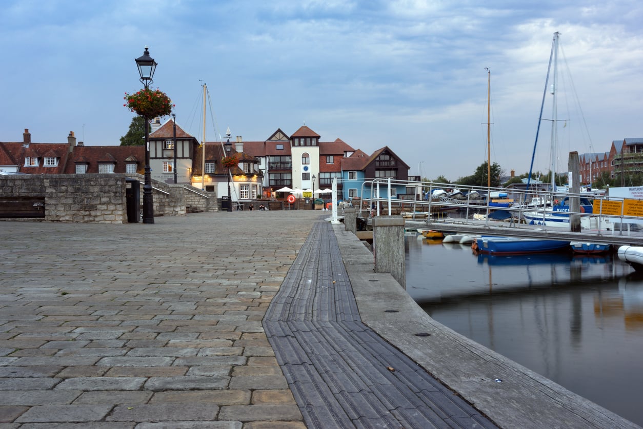 A peaceful harbor scene with moored boats on calm water, a cobblestone walkway, and charming waterfront buildings under a cloudy sky.